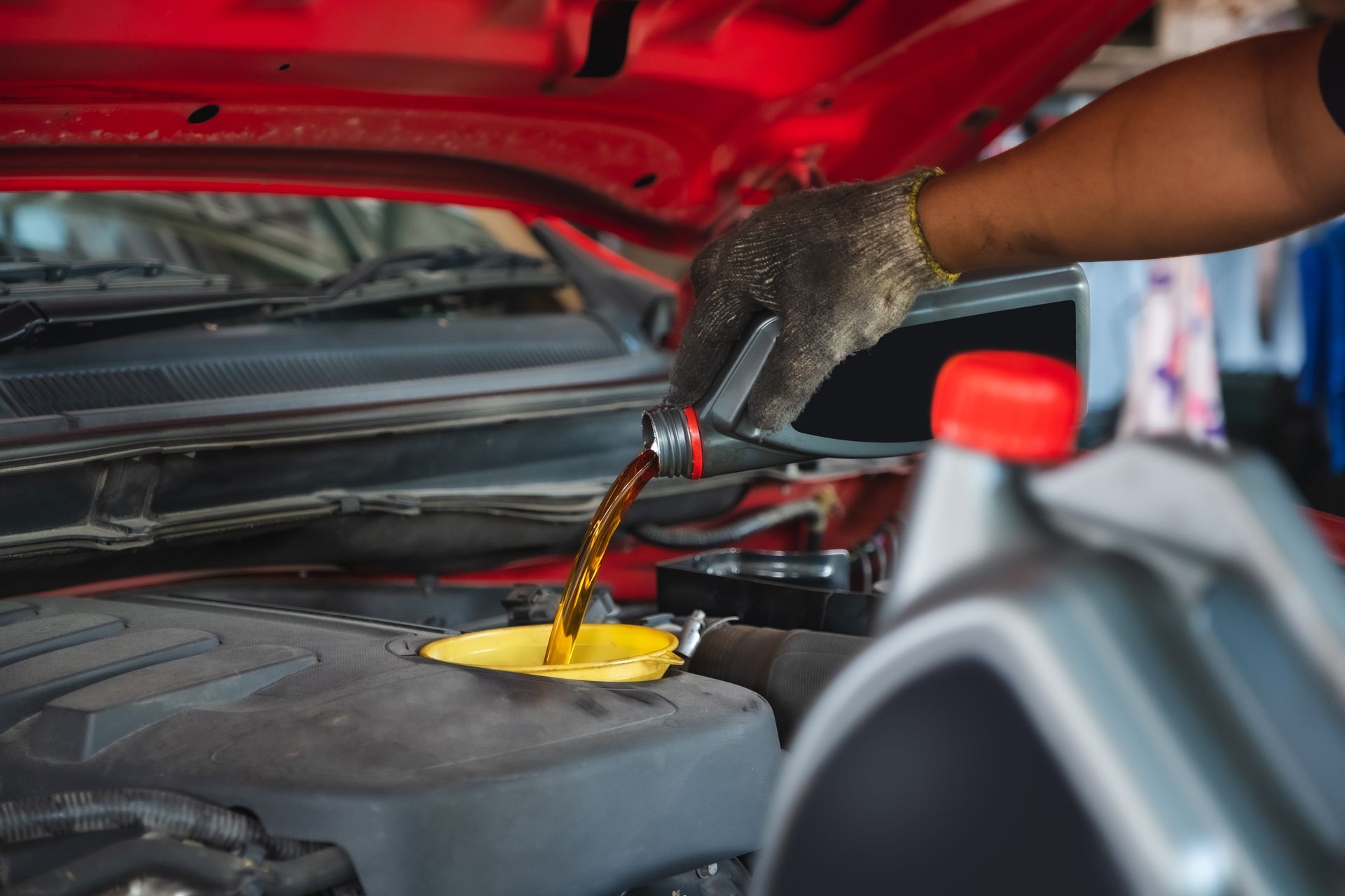 Person pouring oil into a car engine; red car hood open; gloves on hand.