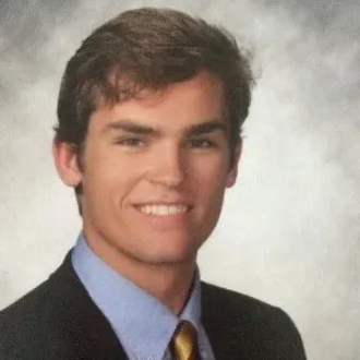 Man in suit and tie smiling at the camera. He has brown hair and is wearing a blue shirt.