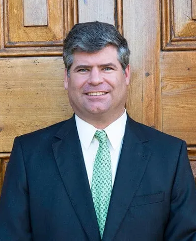 Man in a suit smiles, standing in front of a wooden door. Green tie, dark suit jacket.