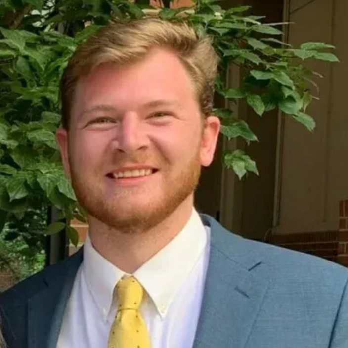 Man in a blue blazer and yellow tie smiles, with leafy green background.