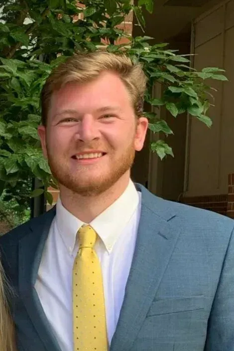Man in a blue suit and yellow tie smiles, outdoors with foliage in the background.