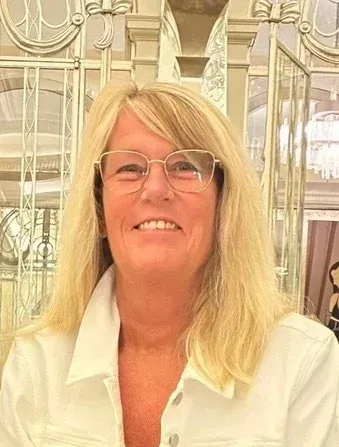 Woman with blonde hair and glasses smiles in front of a decorative background, wearing a white jacket.