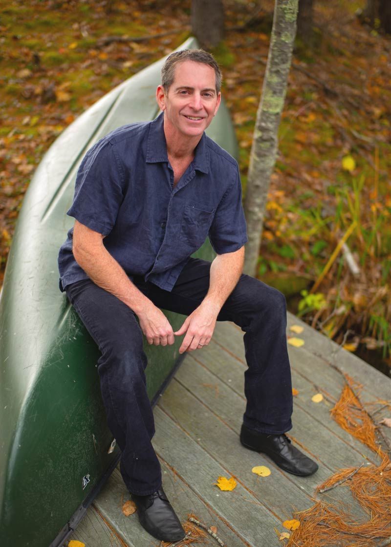 Dr. Louis Granirer is sitting on a tipped green canoe, wearing a blue shirt and dark pants, in a wooded area.