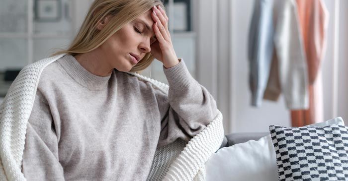 Woman with hand on forehead, wrapped in a blanket, looking unwell, in a living room. Chronic Fatigue Syndrome Remedies - Dr. Louis Granirer Holistic Chiropractor