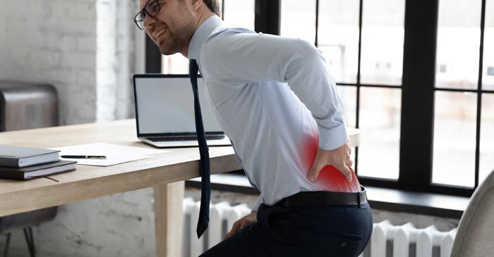A man is clutching his lower back in pain at his desk. Red highlight on pain area indicating Herniated Disc Treatment - Dr. Louis Granirer, Holistic Chiropractor.