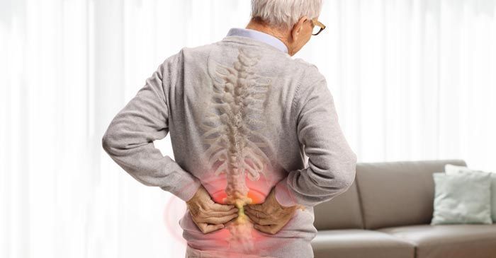 Man with back pain, spine highlighted. He's holding his lower back near a couch needing Lower Back Pain Treatment - Dr. Louis Granirer Holistic Chiropractor