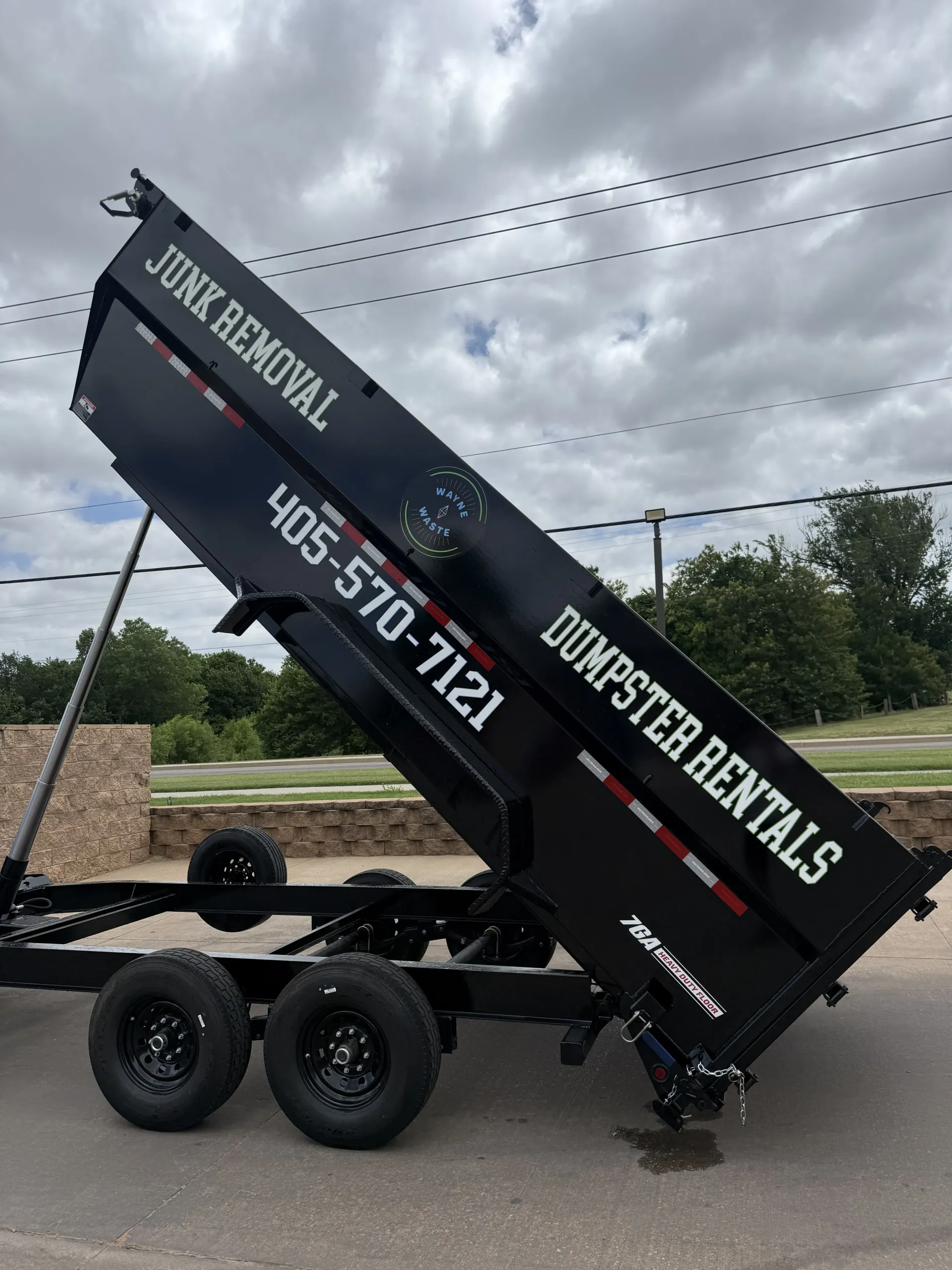Black dump trailer with raised bed, “Dump Trailer Rentals,” parked outdoors under cloudy sky.