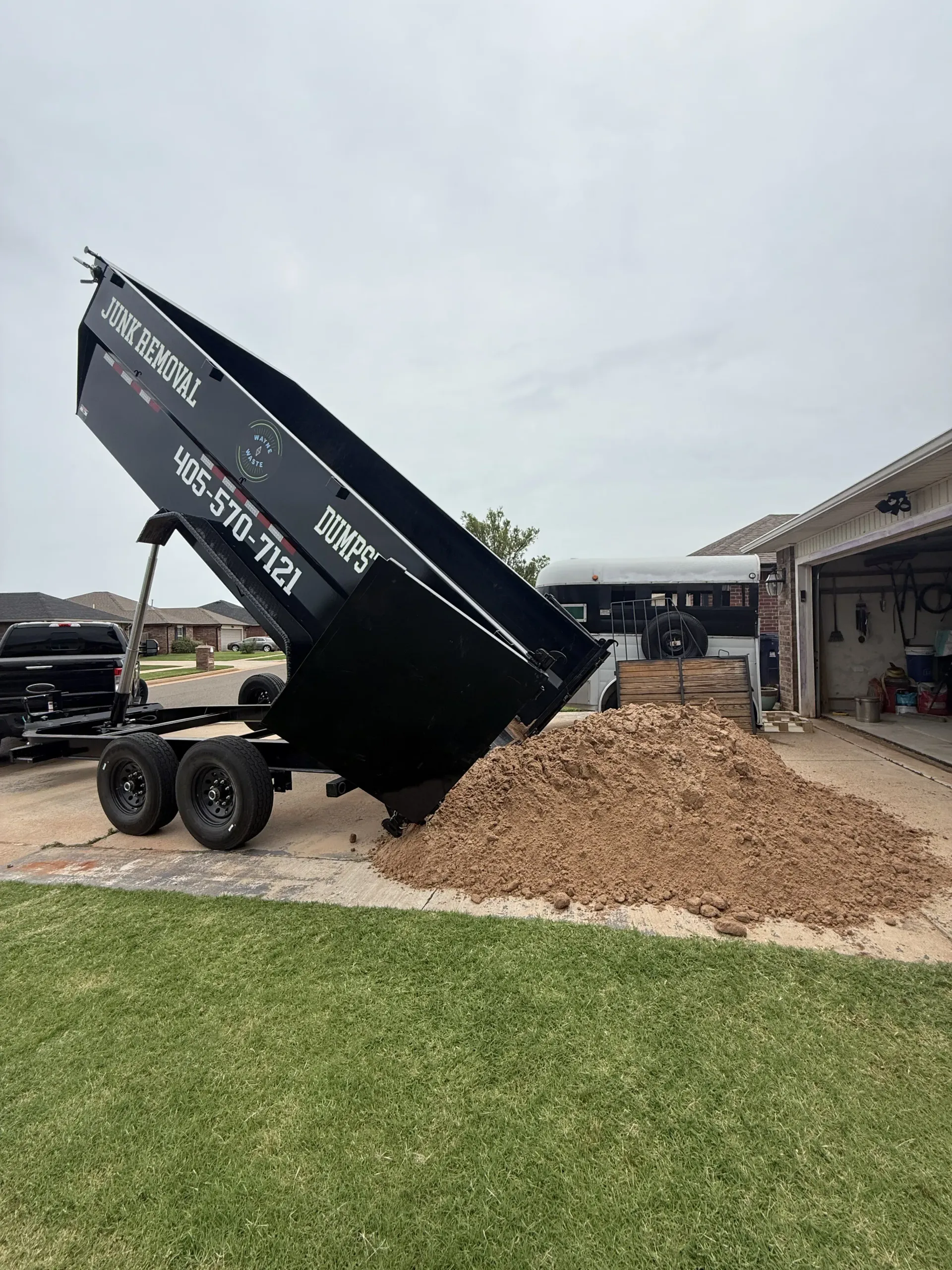 Black dump trailer unloading mulch pile beside a garage and lawn in a suburban driveway