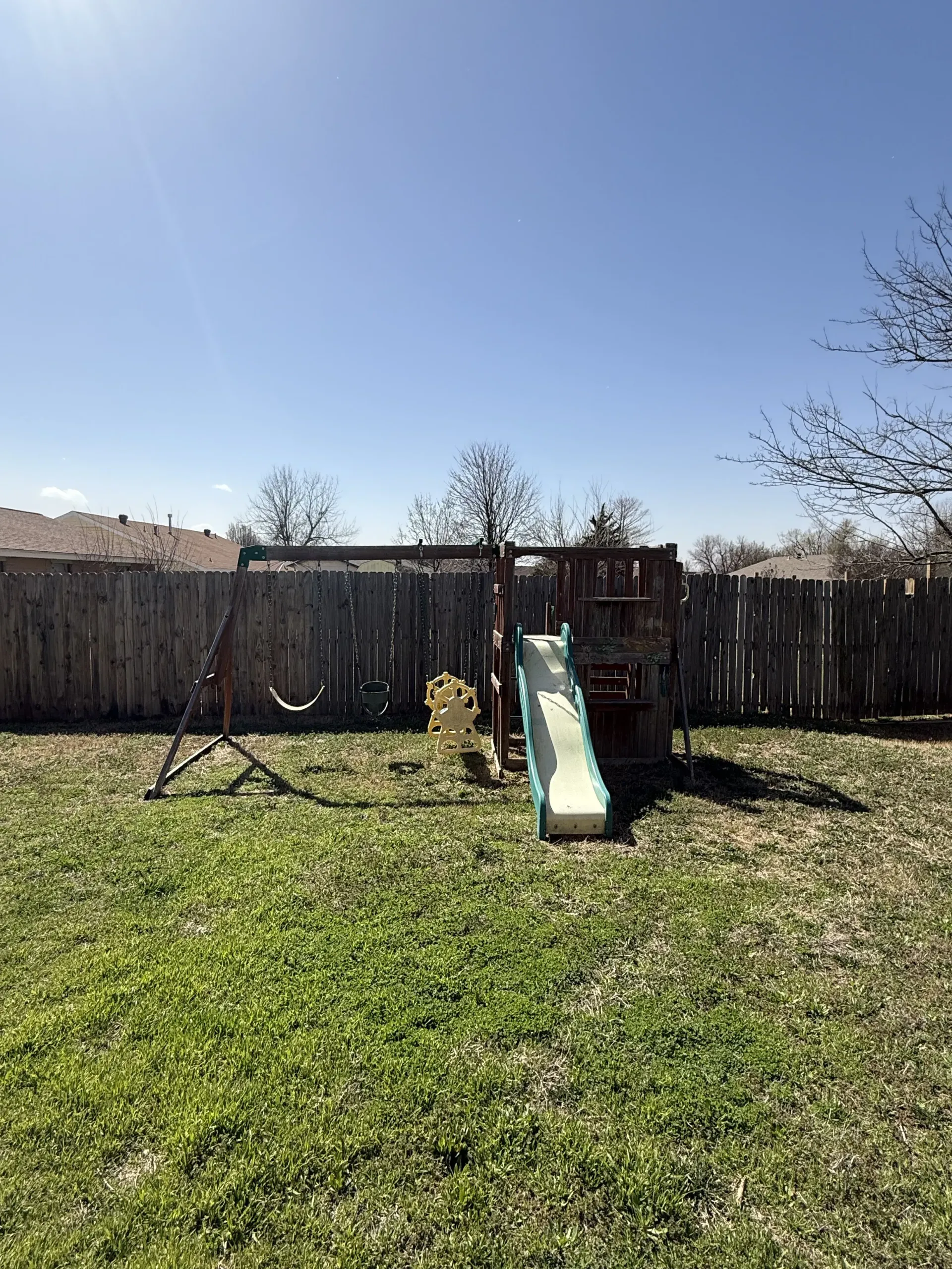 Backyard playground with a slide, fenced yard, and grassy lawn under a clear blue sky