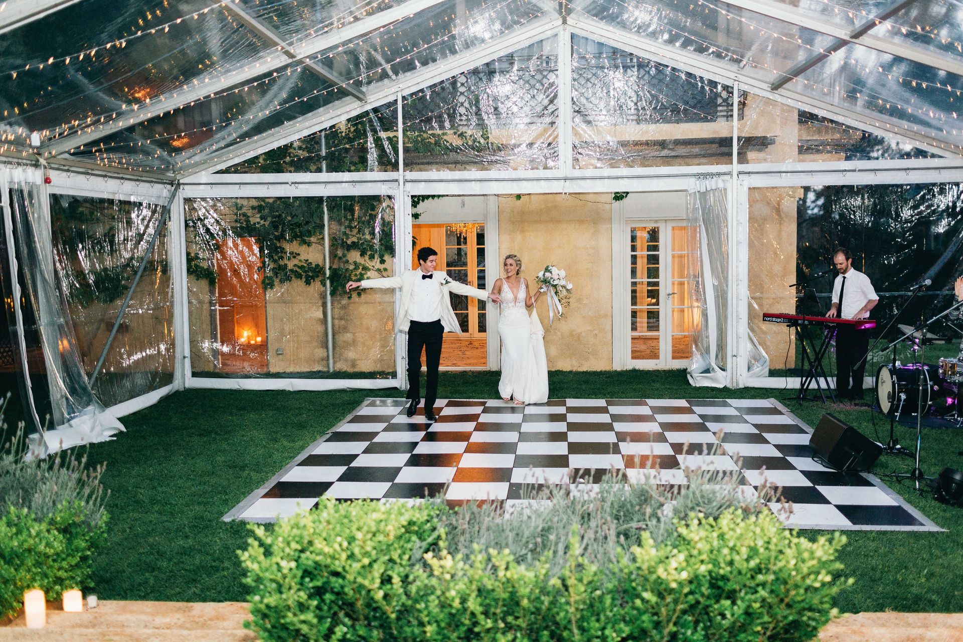 A bride and groom are dancing on a checkered dance floor under a clear tent.