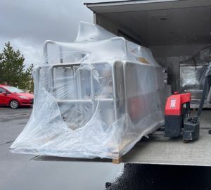 Stór iðnaðarvél vafin inn í plast á bretti, affermd af vörubíl með brettijakki.