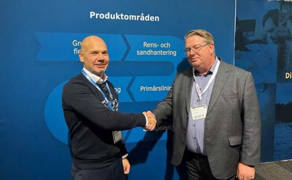 Tveir menn taka í höndina á sér fyrir framan bláan bakgrunn með texta, hugsanlega á viðskiptasýningu eða ráðstefnu.