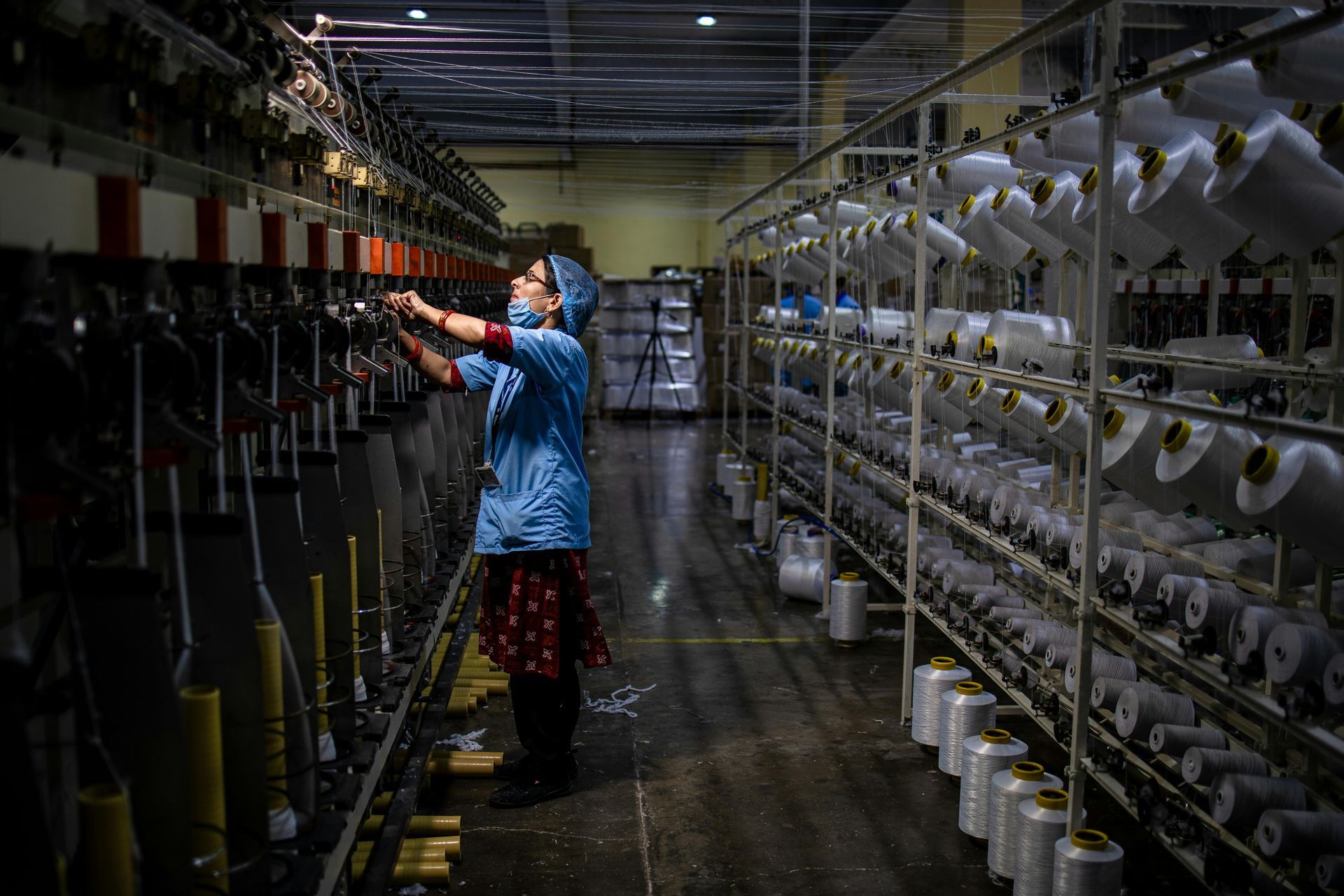 Verkamaður í bláum kyrtil stjórnar vefnaðarvél í verksmiðju fullri af röðum af hvítum þráðspólum.