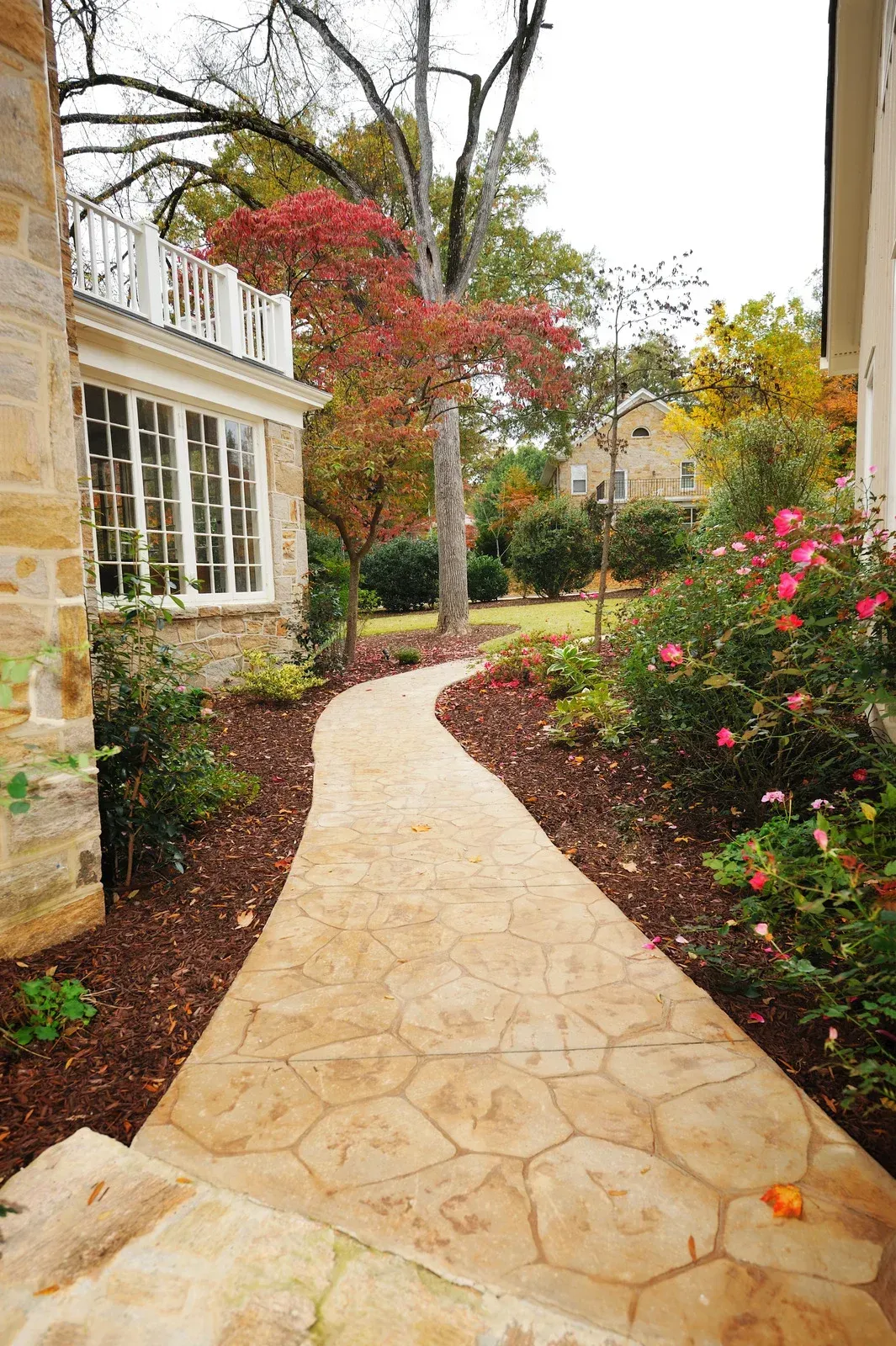 Stone walkway winds through a garden with colorful foliage, leading to a house.