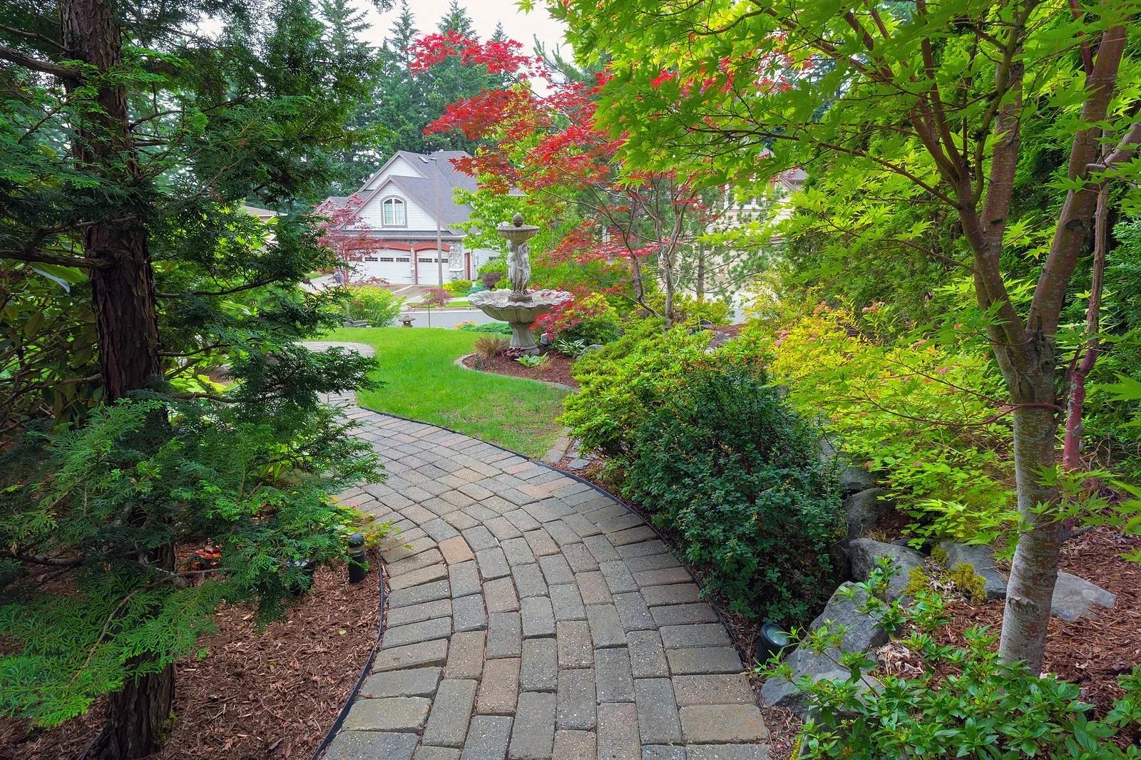 Brick pathway leads to a house, surrounded by lush green and red foliage.