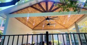 Outdoor patio with wood-paneled ceiling, two ceiling fans, and a black railing, under a white roof.
