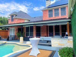 Backyard with a pool, patio, and brick house under a blue sky; a white table stands in the foreground.