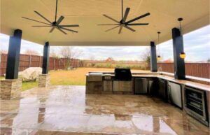 Covered outdoor kitchen with fans, appliances, and seating area overlooking a backyard.