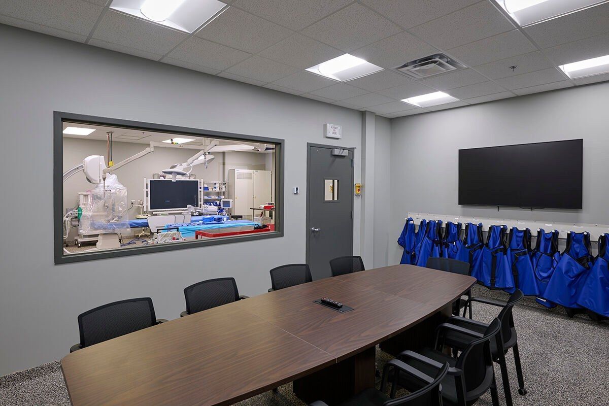 A conference room with a long table and chairs and a large window with a view of an operating room.