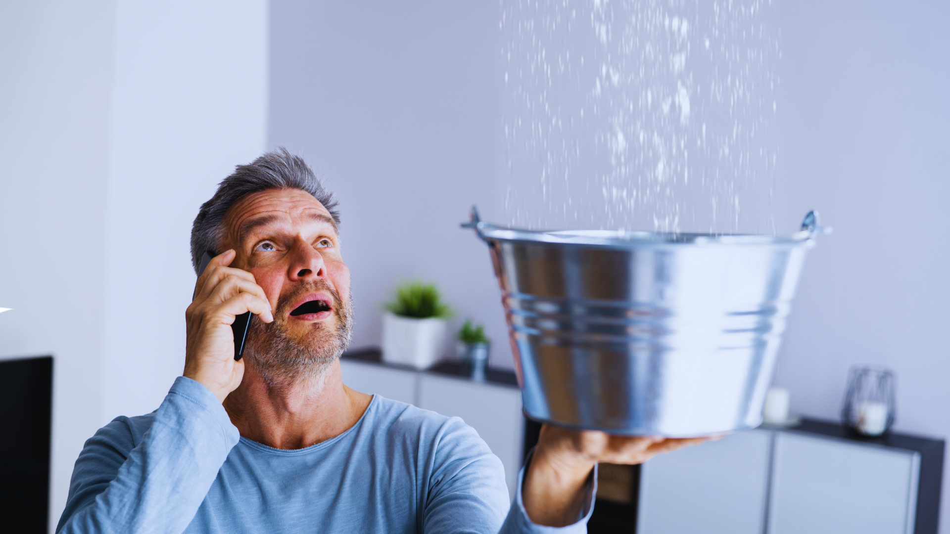 A man is talking on a cell phone while holding a bucket with water coming out of the ceiling.