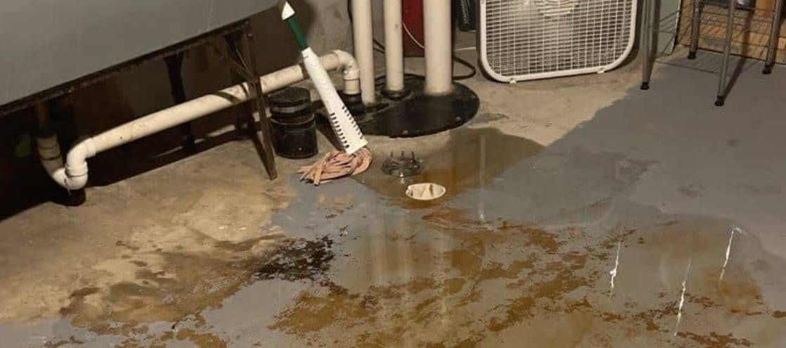A flooded basement with standing water. Pipes and a fan are visible, along with a mop.