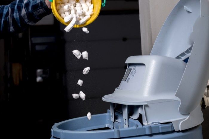 Person pouring salt pellets from a yellow bucket into a water softener unit.