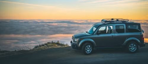 A gray SUV parked on a high mountain overlook, silhouetted against a sea of clouds at sunset.