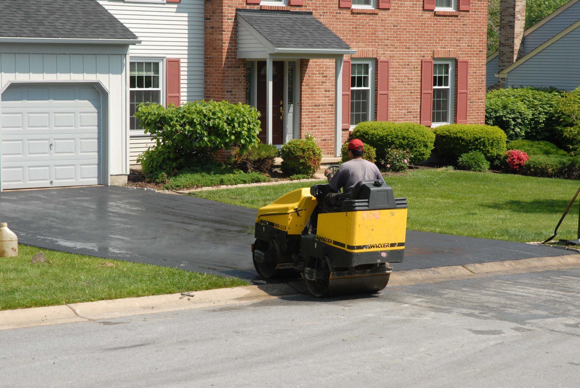 A man is driving a yellow roller on a driveway in front of a brick house