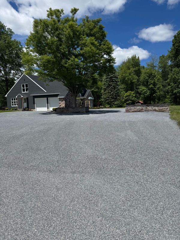 A gravel driveway leading to a house with a tree in the middle.