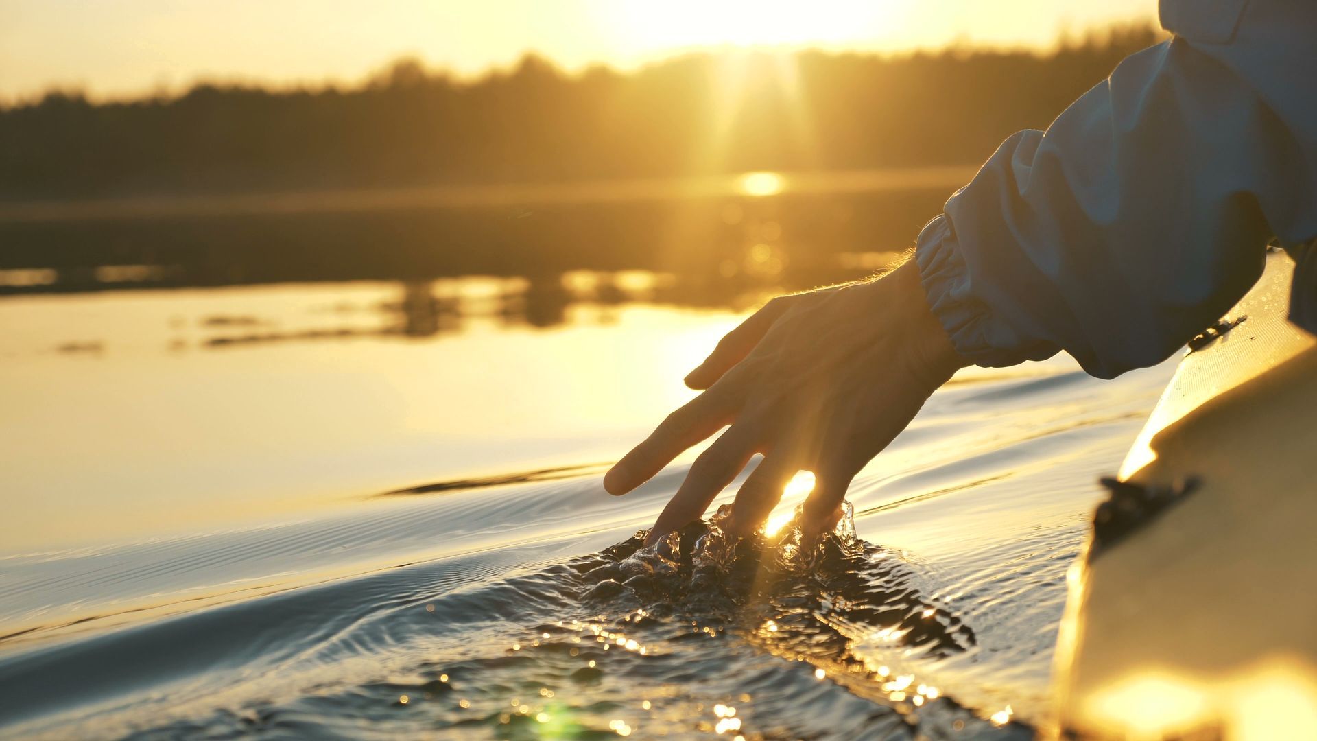 A person’s hand trails through calm lake water at sunset, creating ripples as light reflects off the golden surface.