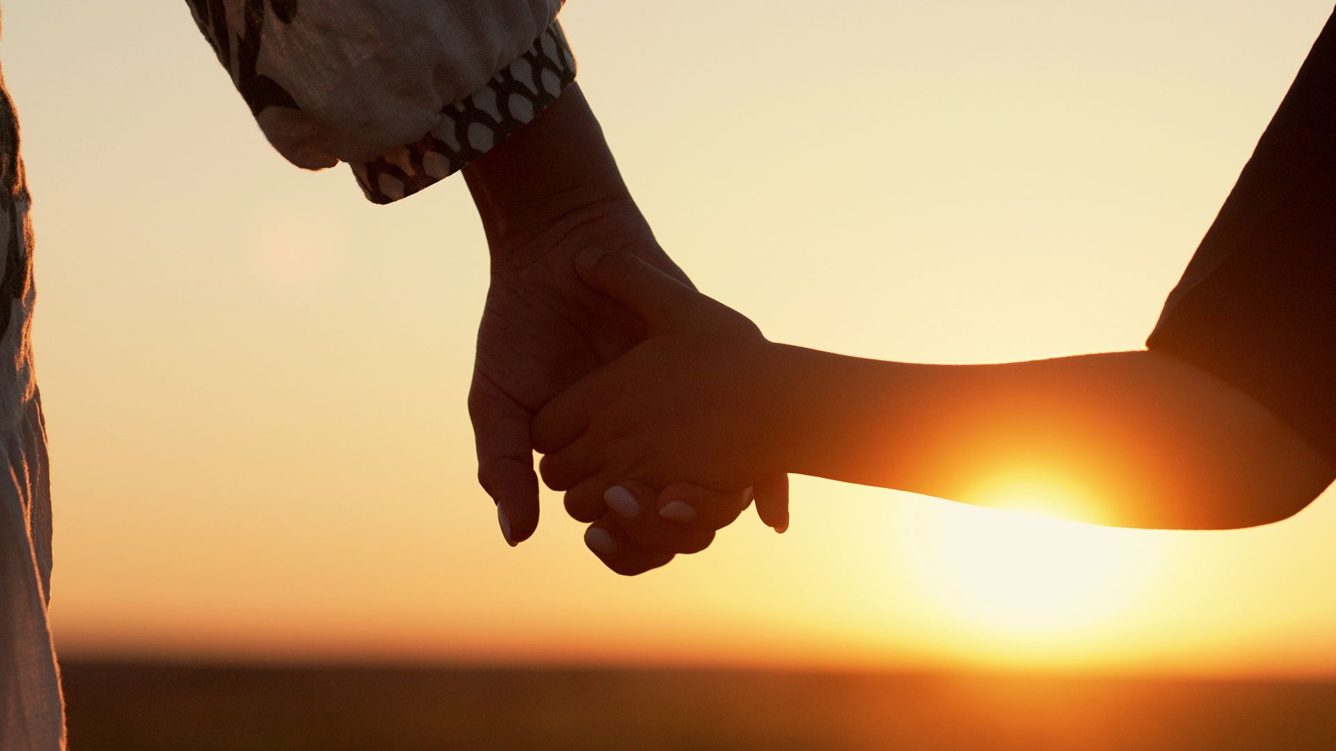 Two people hold hands in silhouette against a warm, glowing sunset.