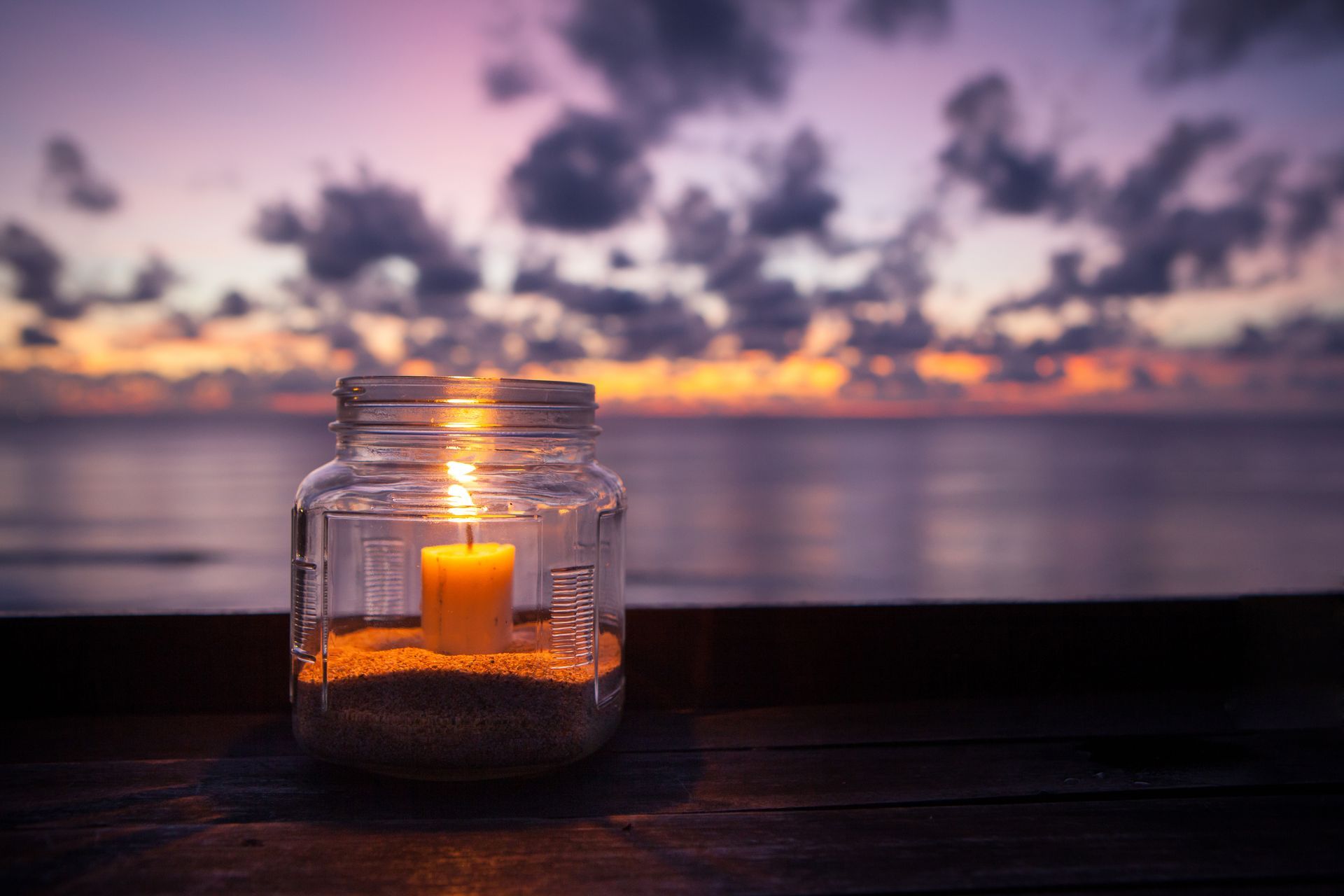 A lit candle sits in a glass jar filled with sand on a wooden surface, with a sunset over the ocean in the background.
