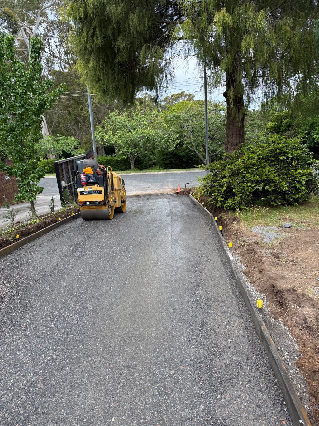 A Yellow Construction Roller Compacts Gravel on A Residential Driveway Lined with Wooden Forms and Yellow Markers — About Time Plumbing & Civil Construction in Moss Vale, NSW