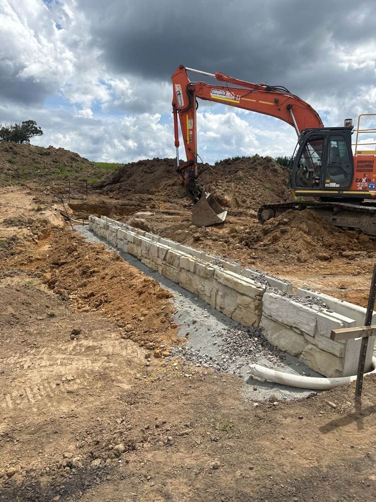 An Excavator Is Digging A Trench In A Dirt Field — About Time Plumbing & Civil Construction in Moss Vale, NSW