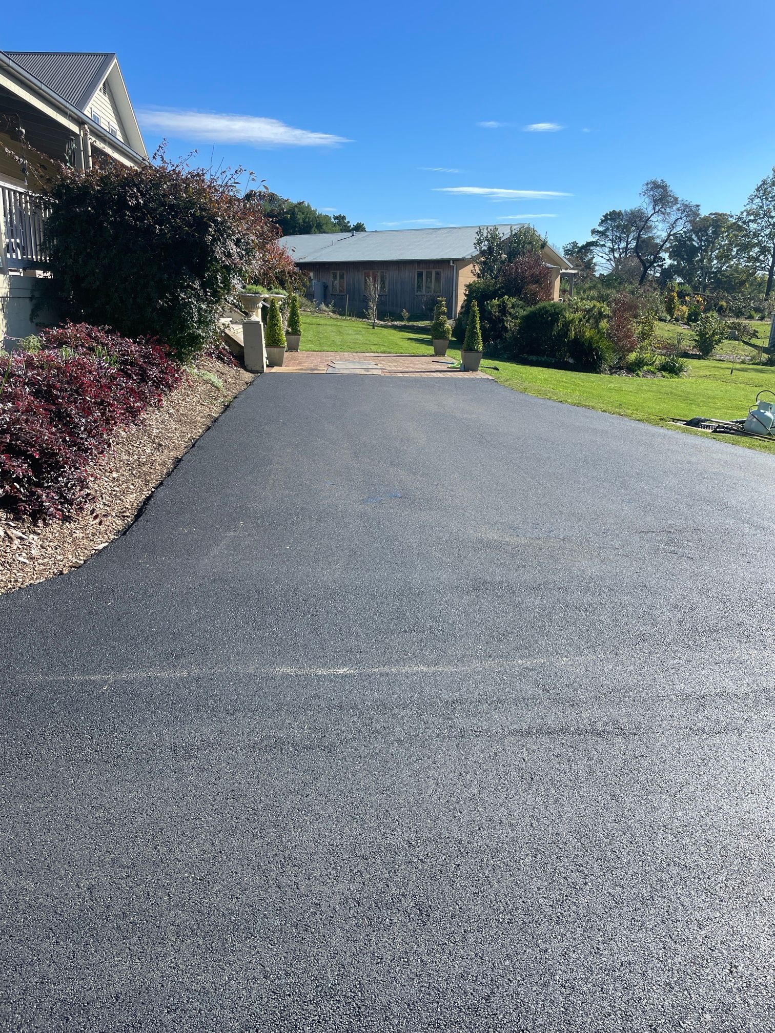 A Black Asphalt Driveway Leading To A House On A Sunny Day — About Time Plumbing & Civil Construction in Moss Vale, NSW