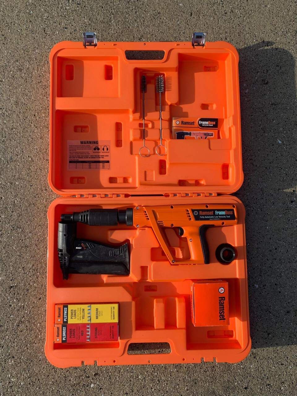 Orange toolbox open, containing a nail gun and accessories on pavement.