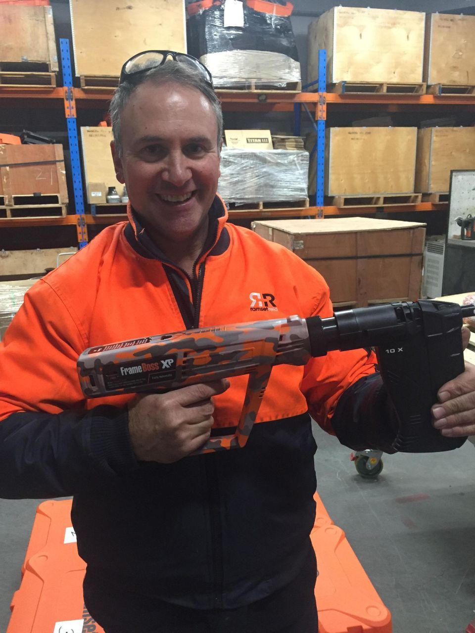 Man smiling, holding a large nail gun. He wears an orange and black work shirt. Warehouse setting.