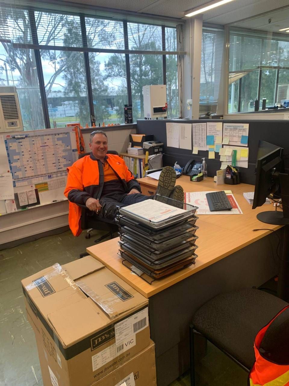Man in orange workwear sits at desk with feet up, looking at camera in office. Several boxes and papers are present.