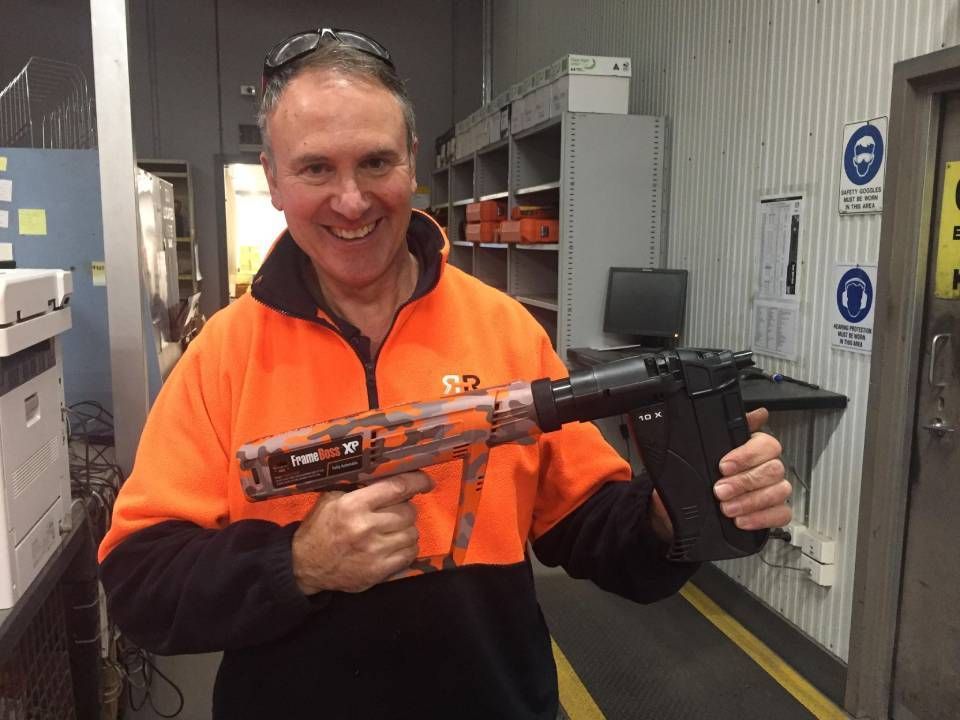 Man in orange workwear holding a powder-actuated nail gun indoors, smiling.