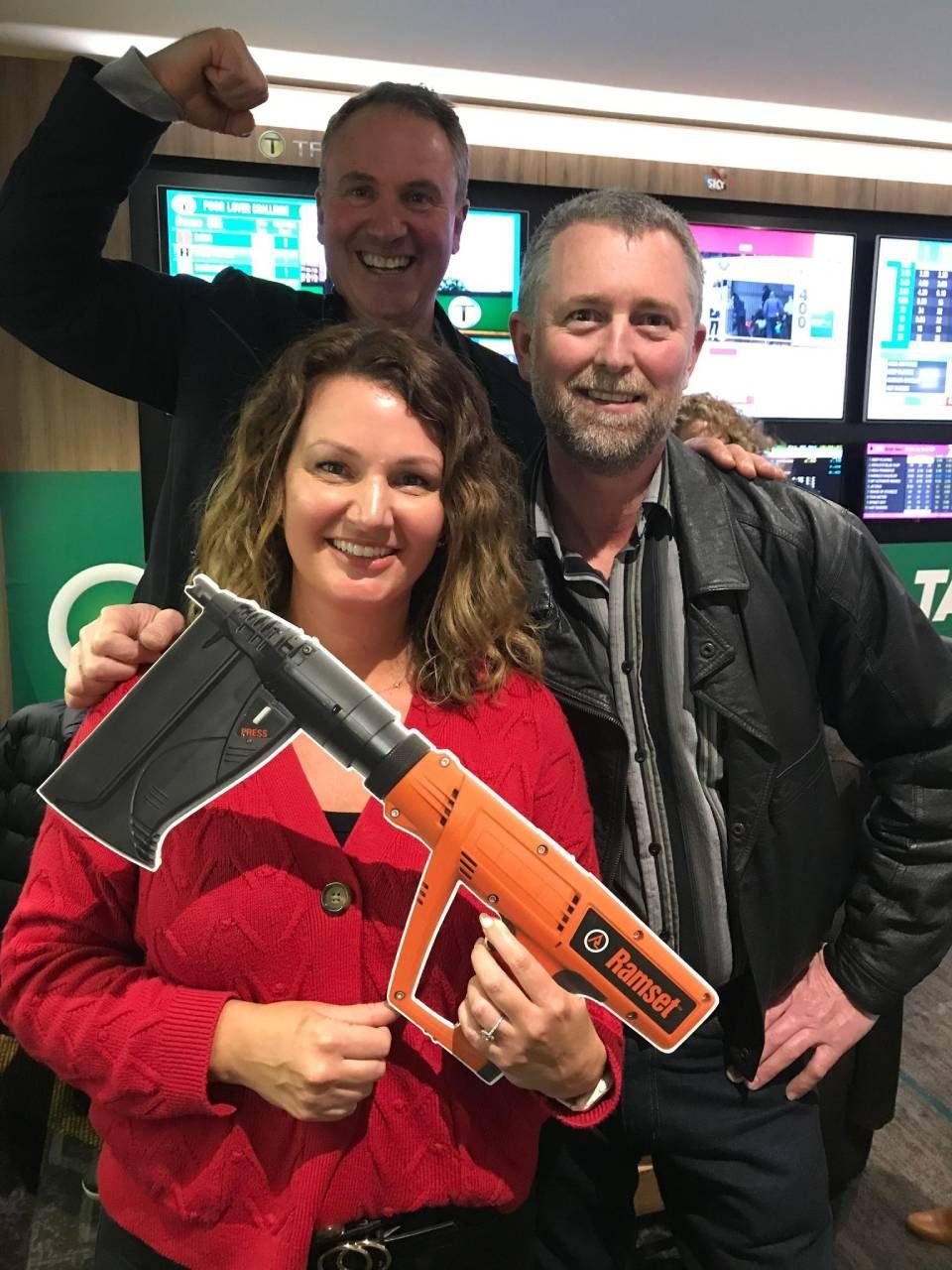 Three people smiling, one holding an orange power tool cutout in a venue with screens.
