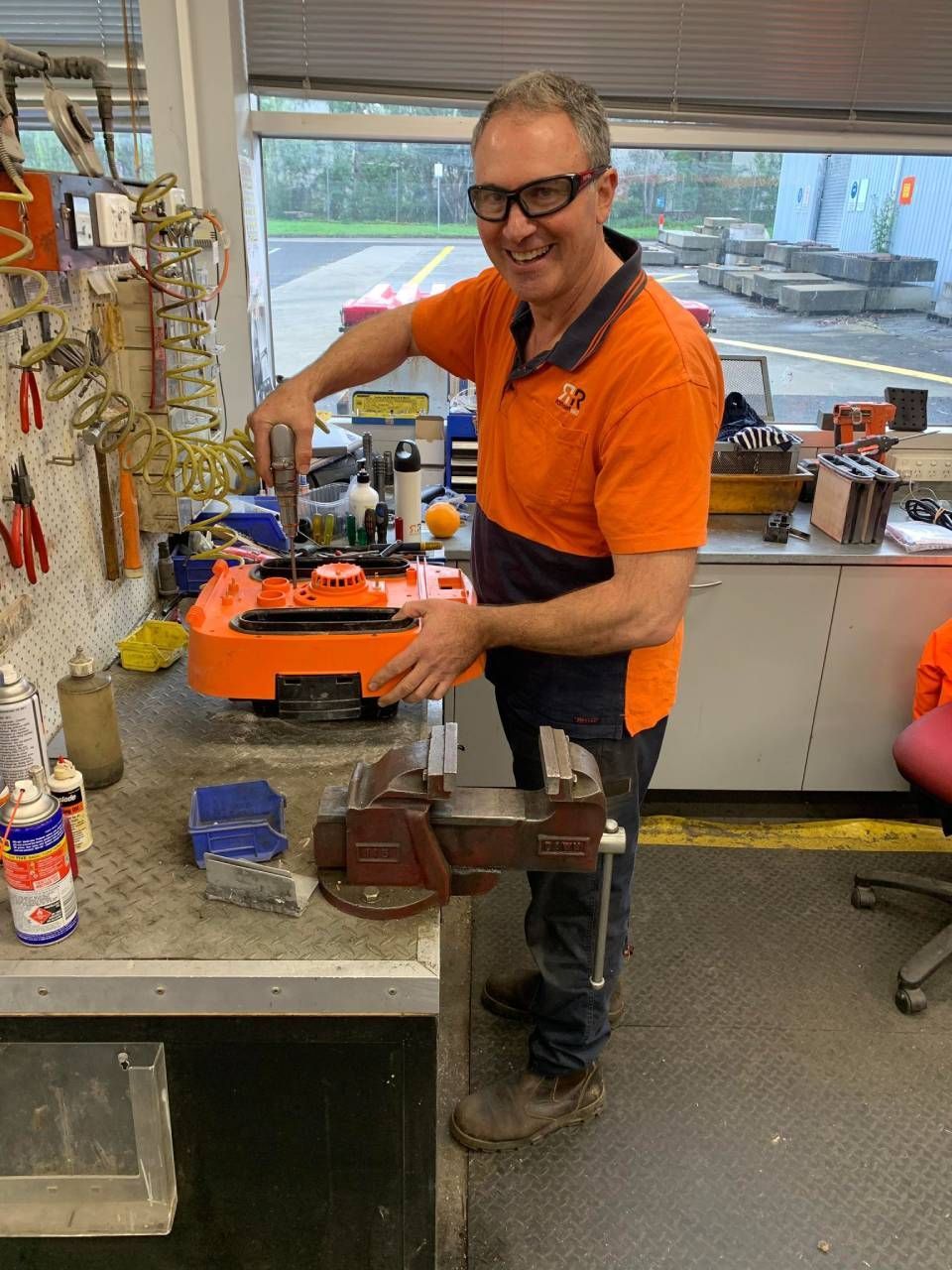 Man in orange shirt works on orange machine at a workbench. He smiles, in workshop setting.