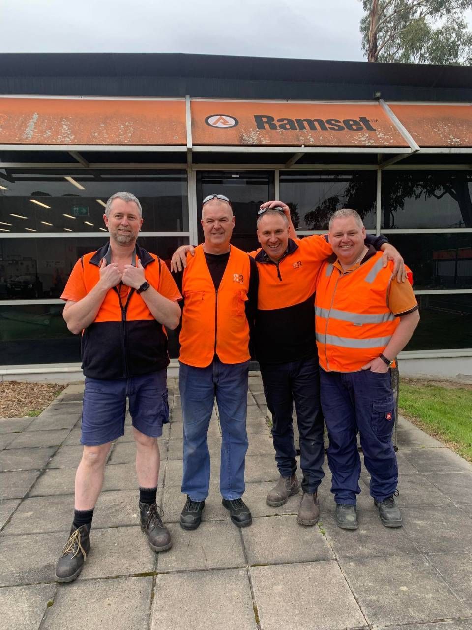 Four men in orange safety vests pose in front of a building with the name 