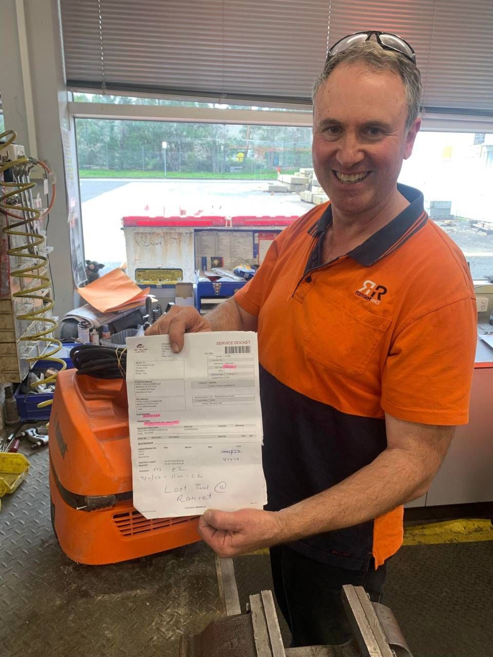 Man in orange shirt smiles, holding a document, in a shop with machinery.