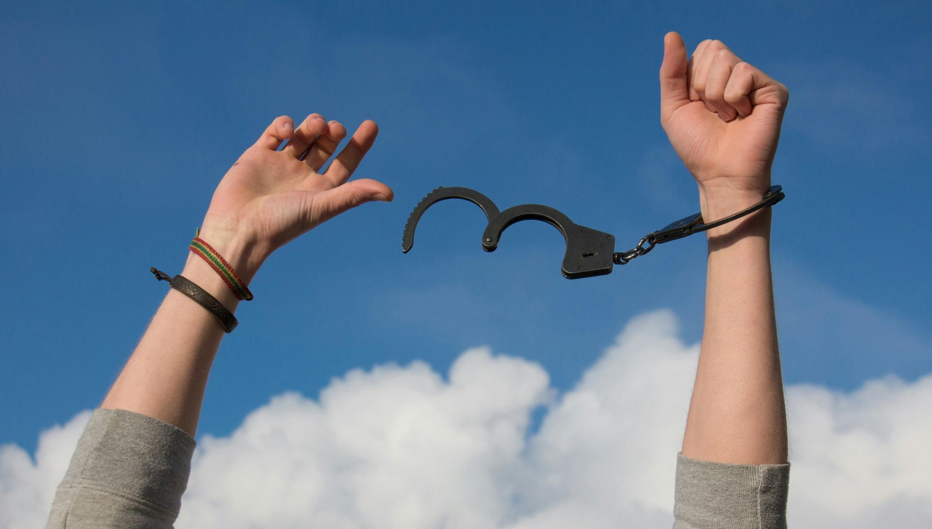 An image of hands breaking free of handcuffs with a blue sky and clouds in the background.