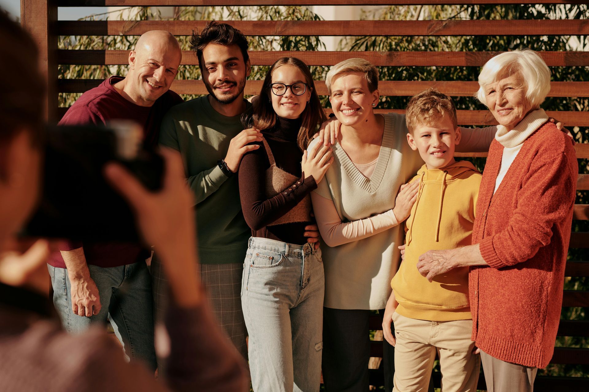 Family poses for a photo outdoors. People smile, arms around each other. Wooden fence in background.
