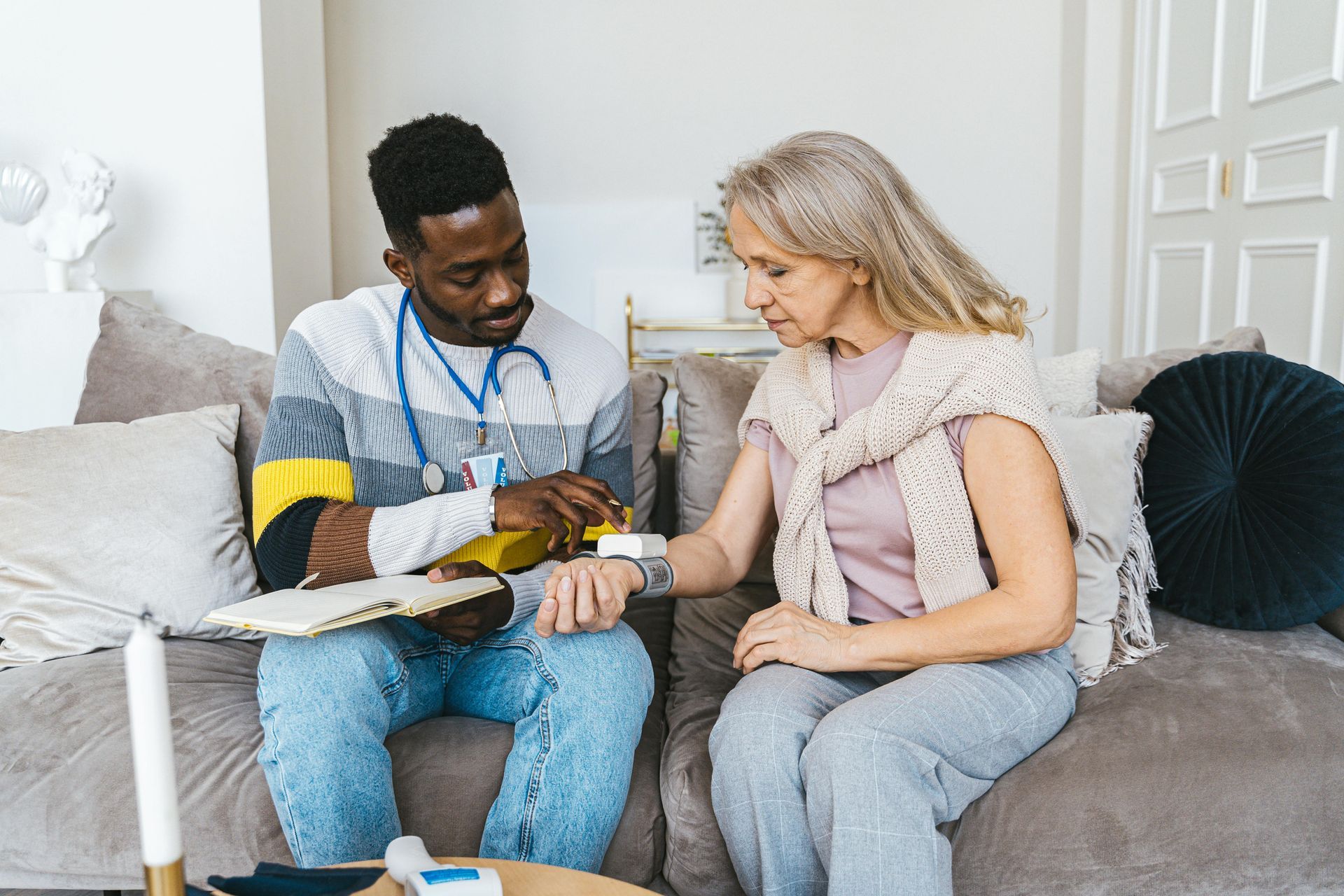 Healthcare worker checks a patient's wrist pulse on a couch. The worker wears a stethoscope and the patient wears a sweater.