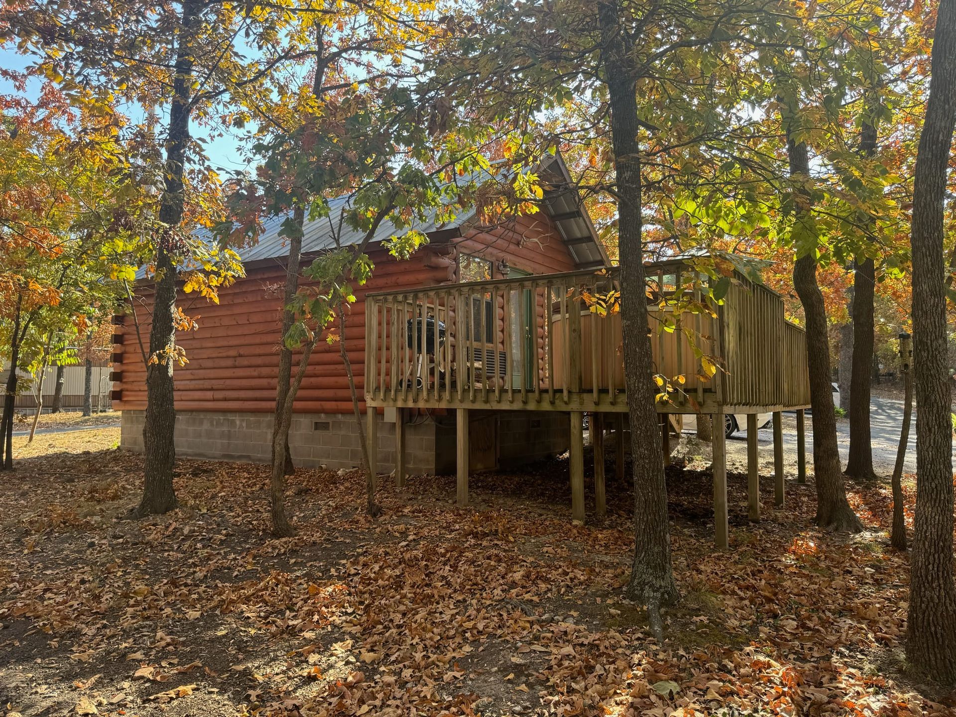 A log cabin with a large deck is surrounded by trees and leaves.