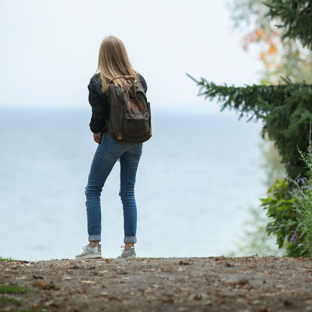 A woman with a backpack is standing on a hill overlooking the ocean.