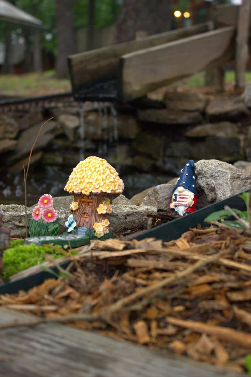 A gnome is sitting on a rock in a fairy garden next to a waterfall.