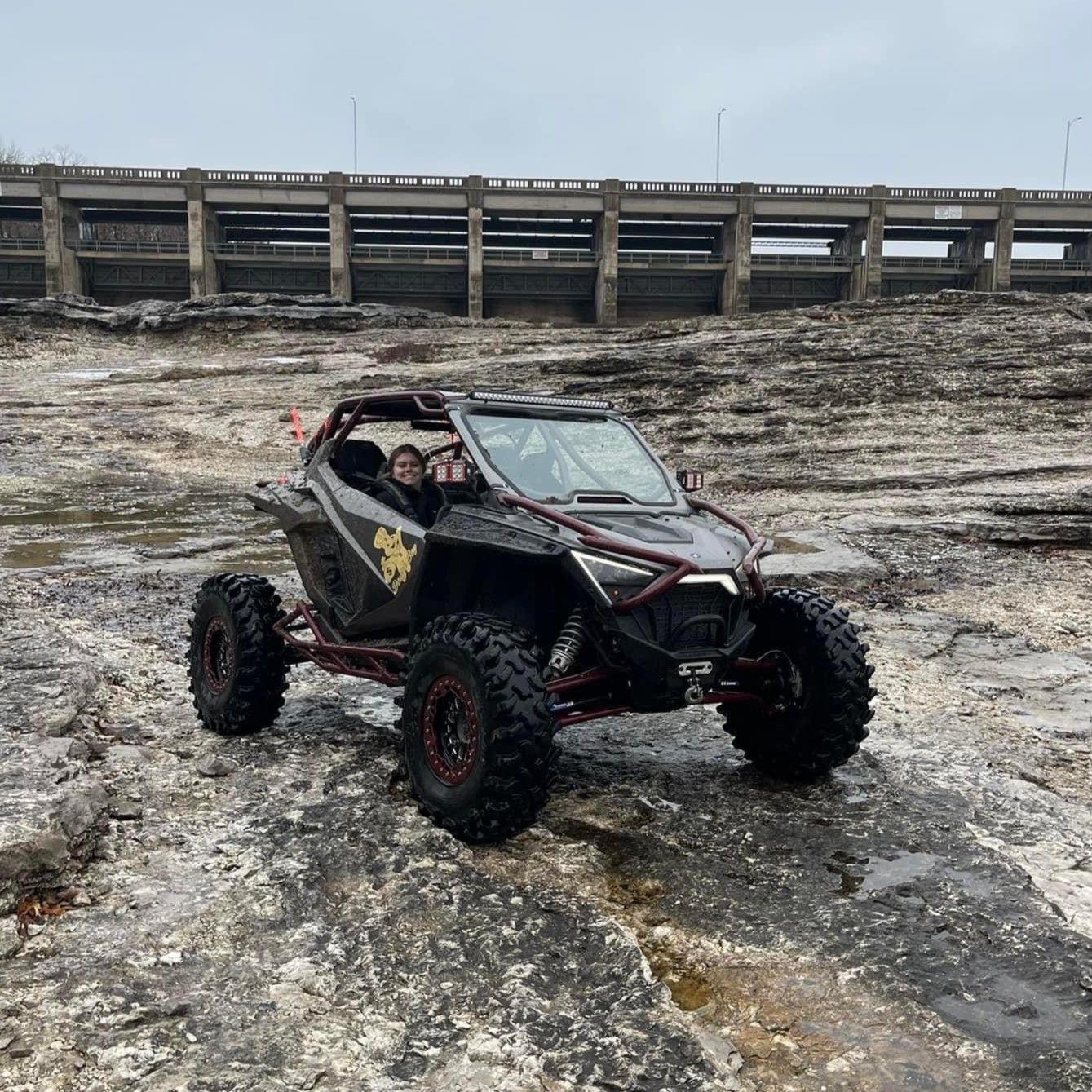 A black atv is driving through a muddy field next to a bridge.